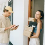 Wanita menerima paket kiriman dari kurir di lorong, menggunakan smartphone untuk konfirmasi pembayaran online